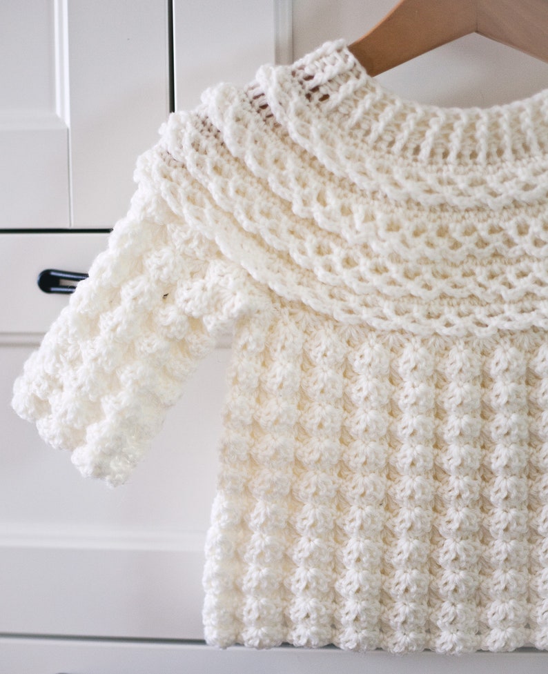 K&ouml;nnte beinhalten: Ein wei&szlig;er H&auml;kelpullover mit einem breiten, gekr&auml;uselten Kragen und einem strukturierten Stichmuster.