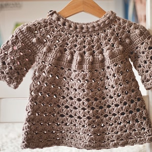Puede incluir: Un jersey de beb&eacute; de punto de ganchillo marr&oacute;n con cuello redondo y mangas cortas. El jersey tiene un patr&oacute;n texturizado con peque&ntilde;as bolitas.