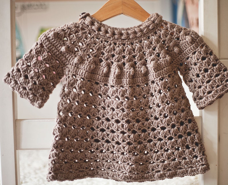 Puede incluir: Un jersey de beb&eacute; de punto de ganchillo marr&oacute;n con cuello redondo y mangas cortas. El jersey tiene un patr&oacute;n texturizado con peque&ntilde;as bolitas.