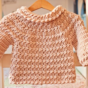 May include: A light pink, hand-crocheted baby sweater with a textured stitch pattern. The sweater features a ruffled collar and cuffs, and is displayed on a wooden hanger. The soft, warm garment is perfect for a baby.