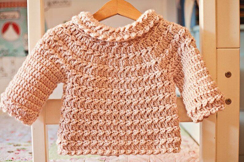 May include: A light pink, hand-crocheted baby sweater with a textured stitch pattern. The sweater features a ruffled collar and cuffs, and is displayed on a wooden hanger. The soft, warm garment is perfect for a baby.