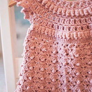 Peut inclure: Une robe de b&eacute;b&eacute; en crochet rose avec un design d&eacute;licat et ajour&eacute; et un ourlet festonn&eacute;.