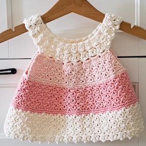 May include: A crocheted baby dress with a pink and white ombre design. The dress has a scalloped hem and a crocheted flower detail around the neckline.