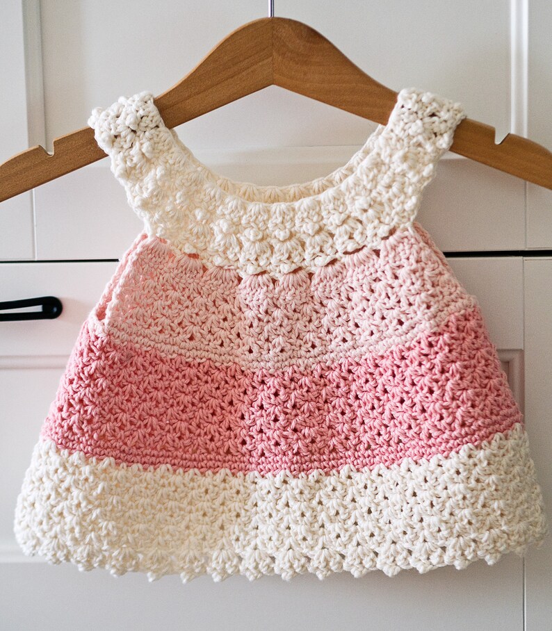 May include: A crocheted baby dress with a pink and white ombre design. The dress has a scalloped hem and a crocheted flower detail around the neckline.