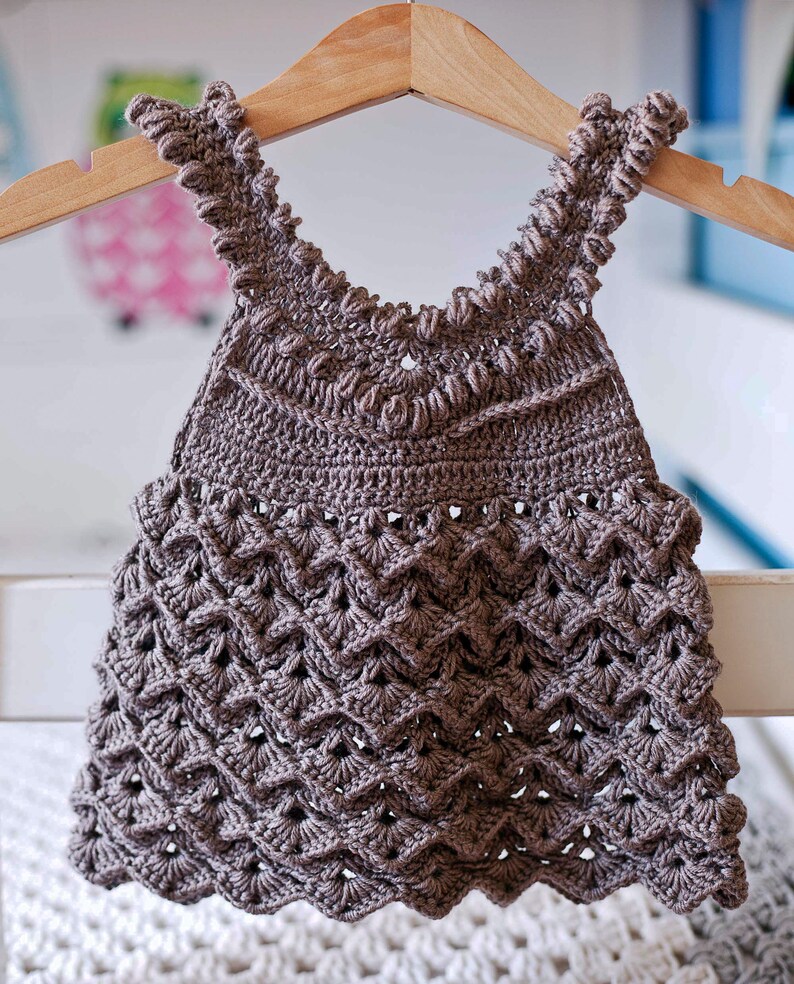May include: A brown crocheted baby dress with a scalloped hem and a delicate crocheted pattern. The dress is hanging on a wooden hanger.