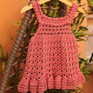 May include: A pink crocheted baby dress with a scalloped hem and straps. The dress is hanging on a wooden hanger.