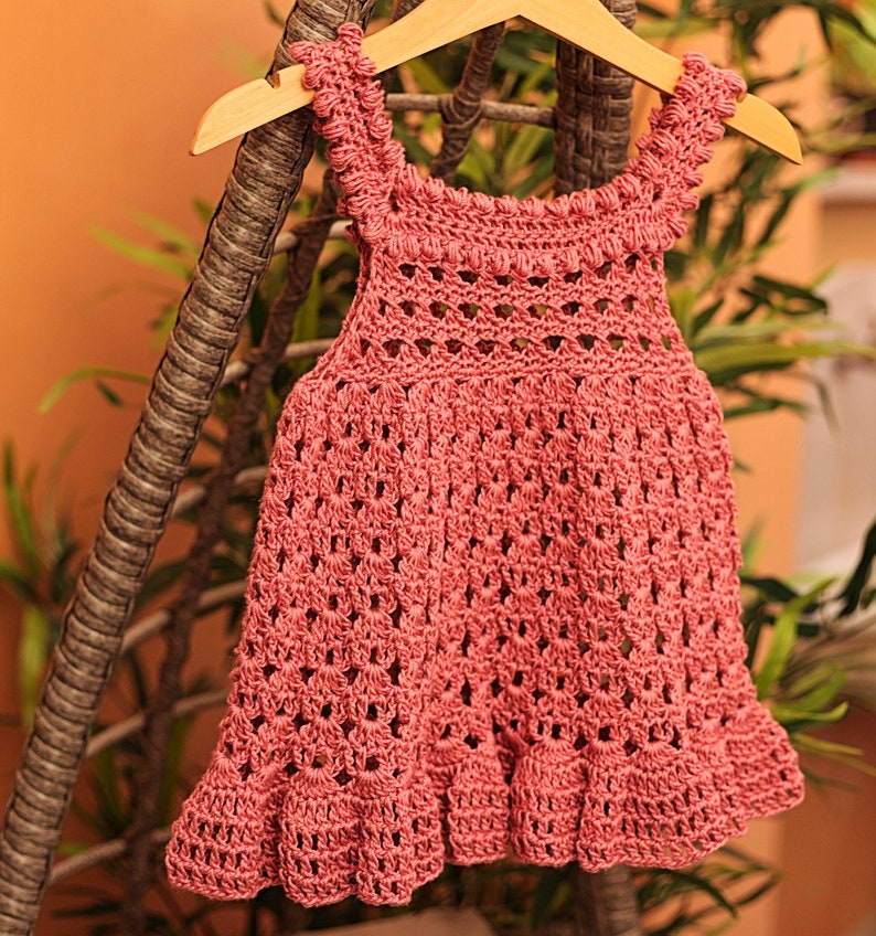 May include: A pink crocheted baby dress with a scalloped hem and straps. The dress is hanging on a wooden hanger.