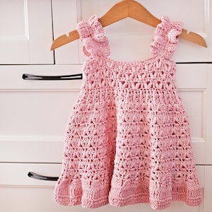 May include: A light pink sleeveless dress crafted with a textured crochet pattern. The garment features ruffled shoulder straps and a flared skirt silhouette. The dress is displayed on a wooden hanger, showcasing the intricate yarn stitch details.