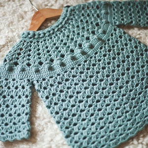 Puede incluir: Un su&eacute;ter de crochet azul claro con un patr&oacute;n delicado. El su&eacute;ter tiene mangas largas y un cuello redondo.