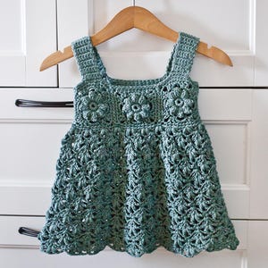 Puede incluir: Un vestido de crochet de color turquesa con un dobladillo festoneado y detalles florales. El vestido tiene un escote cuadrado y tirantes finos.