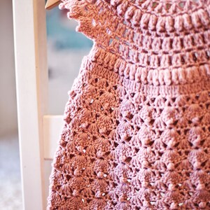 Peut inclure: Une robe de b&eacute;b&eacute; en crochet rose avec un motif d&eacute;licat et ajour&eacute;. La robe a un col rond et des manches courtes.