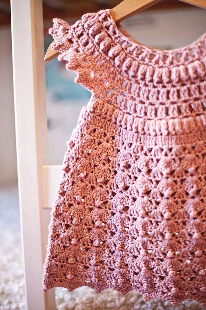 Peut inclure: Une robe de b&eacute;b&eacute; en crochet rose avec un motif d&eacute;licat et ajour&eacute;. La robe a un col rond et des manches courtes.
