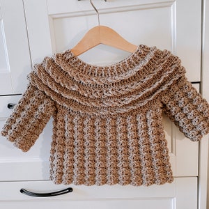 K&ouml;nnte beinhalten: Ein brauner H&auml;kelpullover mit einem R&uuml;schenkragen und einem strukturierten Muster. Der Pullover hat lange &Auml;rmel und eine lockere Passform.
