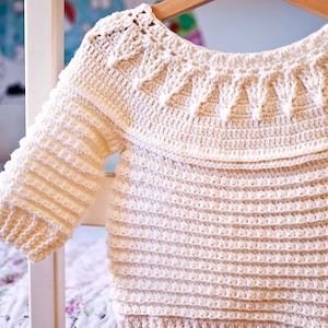 May include: Cream-colored, handmade crochet sweater with long sleeves. The sweater features a detailed, textured pattern around the neckline and on the sleeves. The body of the sweater has a horizontal ribbed design. The sweater is hanging on a wooden hanger.