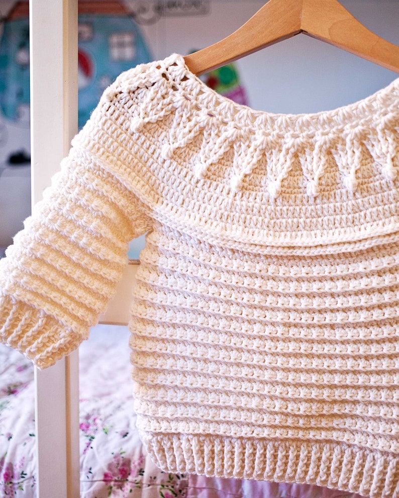 May include: Cream-colored, handmade crochet sweater with long sleeves. The sweater features a detailed, textured pattern around the neckline and on the sleeves. The body of the sweater has a horizontal ribbed design. The sweater is hanging on a wooden hanger.