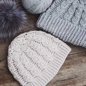 May include: Two knitted hats, one in a light beige color and the other in a light gray color, with a cable knit pattern. The hats are lying on a wooden surface.