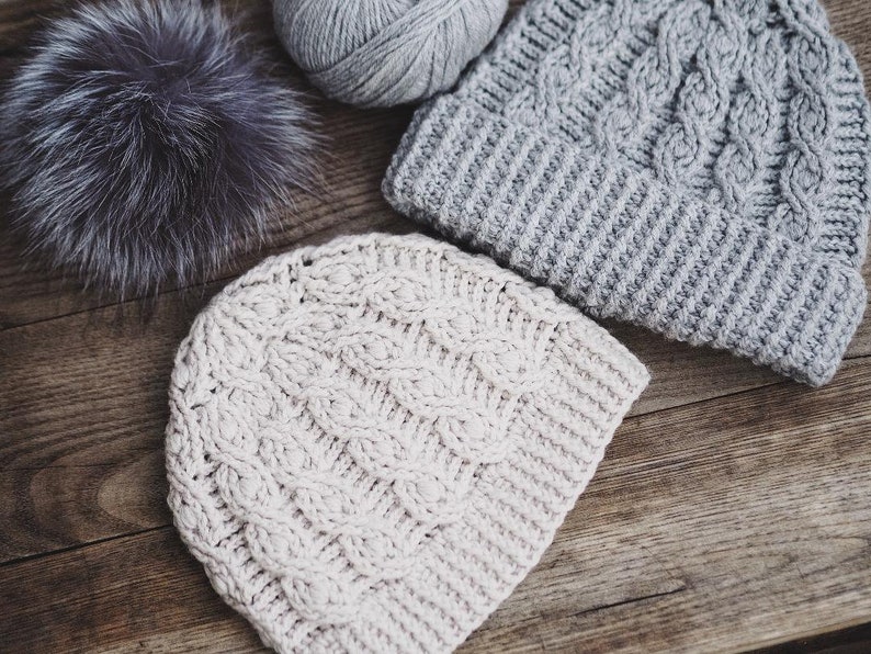 May include: Two knitted hats, one in a light beige color and the other in a light gray color, with a cable knit pattern. The hats are lying on a wooden surface.