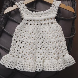 May include: White crocheted baby dress with a scalloped hem and a delicate pattern. The dress is hanging on a wooden hanger.