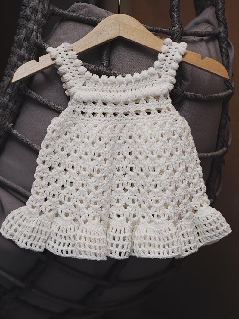 May include: White crocheted baby dress with a scalloped hem and a delicate pattern. The dress is hanging on a wooden hanger.
