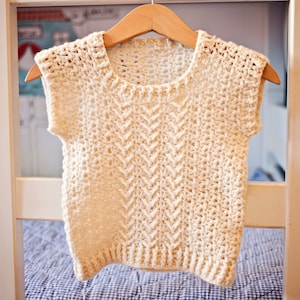 May include: A crocheted sleeveless top with a chevron pattern in a cream color. The top is hanging on a wooden hanger.