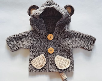 May include: A dark gray crocheted hooded jacket featuring two light beige pockets, two round wooden buttons down the center, and small rounded ears attached to the hood. A small, striped, crocheted tail extends from the bottom hem.