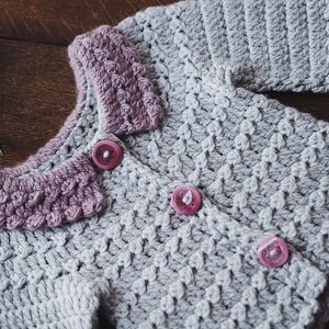 May include: A handmade, light gray crochet cardigan with a textured stitch pattern. The collar and button details are a muted purple. The cardigan has three pink buttons and is displayed on a wooden surface.