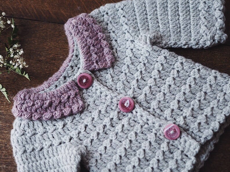 May include: A handmade, light gray crochet cardigan with a textured stitch pattern. The collar and button details are a muted purple. The cardigan has three pink buttons and is displayed on a wooden surface.