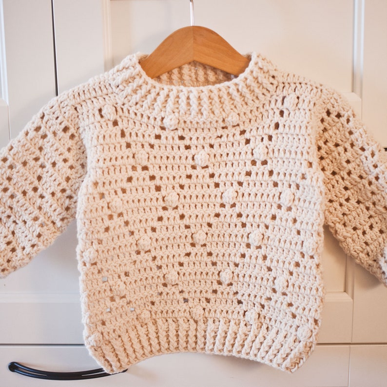 May include: A cream-colored crocheted sweater with a textured stitch pattern and a round neckline. The sweater is hanging on a wooden hanger.
