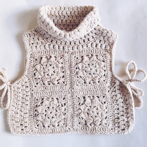 Può includere: Un gilet all'uncinetto color beige con collo alto e motivo a quadrati. Il capo è realizzato con quattro grandi quadrati all'uncinetto con motivi floreali e presenta sottili laccetti laterali per la chiusura.