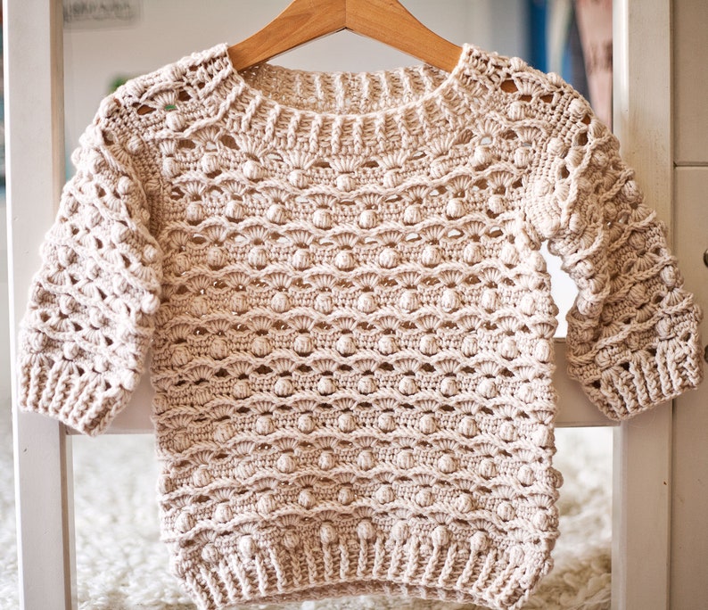 May include: A beige crocheted sweater with a textured, bobble stitch pattern. The sweater has a round neckline and long sleeves.