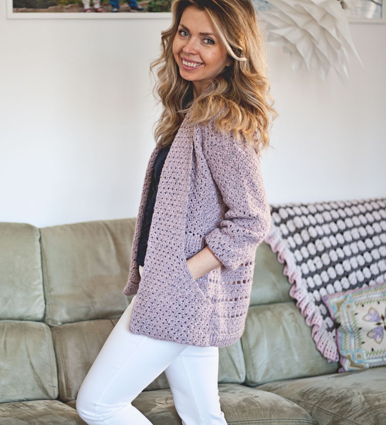 May include: A light purple crocheted cardigan with a loose fit and long sleeves. The cardigan has a V-neck and two pockets. The model is wearing white pants and a dark blue shirt.