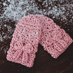May include: A pair of pink crocheted mittens with a bow detail. The mittens are lying on a brown wooden surface covered in white snow.
