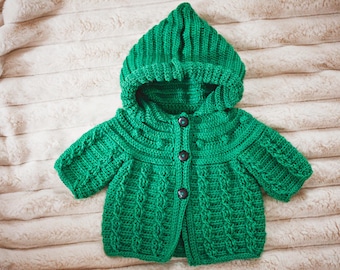 May include: A vibrant green, handmade crochet baby jacket with a hood. The jacket features a button-up front and intricate cable knit detailing. The garment is displayed on a soft, textured surface, showcasing the craftsmanship of the knitted design.