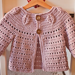 May include: A light pink crocheted cardigan with a button closure. The cardigan has a delicate open stitch pattern and is perfect for a baby or toddler.