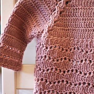 May include: A crocheted sweater in a dusty rose color with a delicate stitch pattern. The sweater has a round neckline and long sleeves.