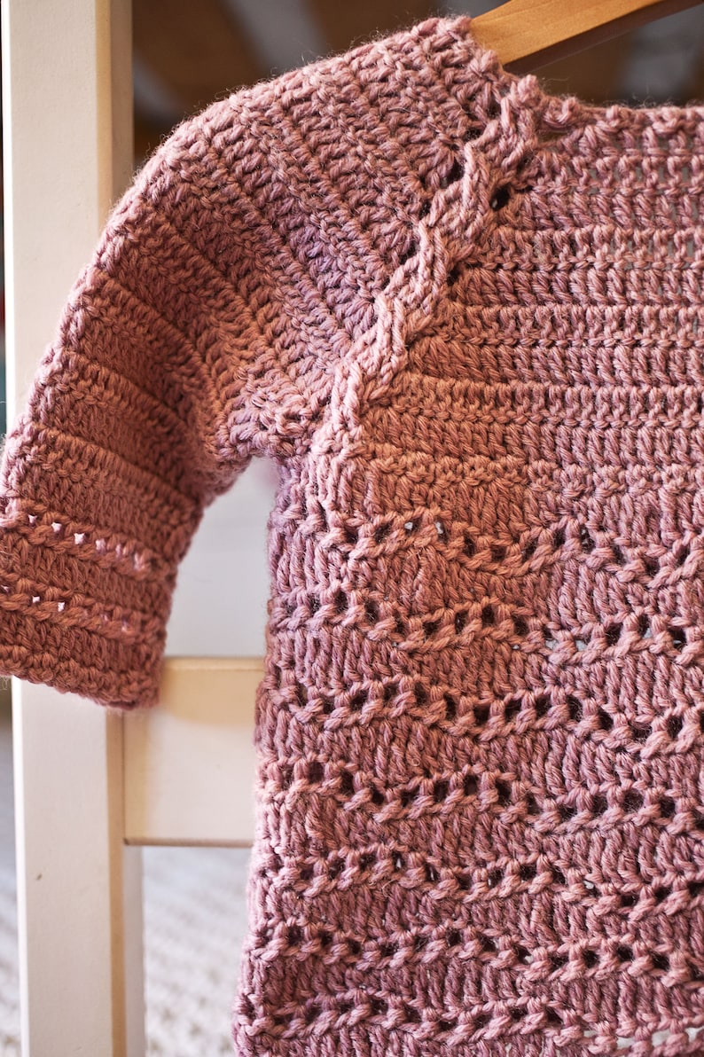 May include: A crocheted sweater in a dusty rose color with a delicate stitch pattern. The sweater has a round neckline and long sleeves.