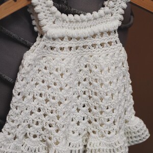 May include: A white crocheted baby dress with a scalloped hem and a delicate crochet pattern. The dress is hanging on a wooden hanger.