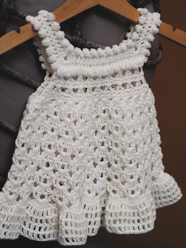 May include: A white crocheted baby dress with a scalloped hem and a delicate crochet pattern. The dress is hanging on a wooden hanger.