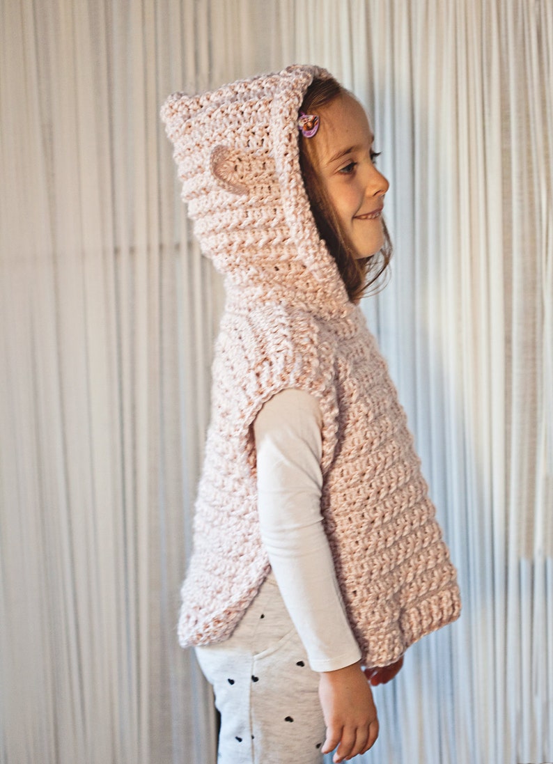 K&ouml;nnte beinhalten: Eine rosa gestrickte Weste mit Kapuze und einem herzf&ouml;rmigen Detail auf der Kapuze. Die Weste hat kurze &Auml;rmel und eine lockere Passform.