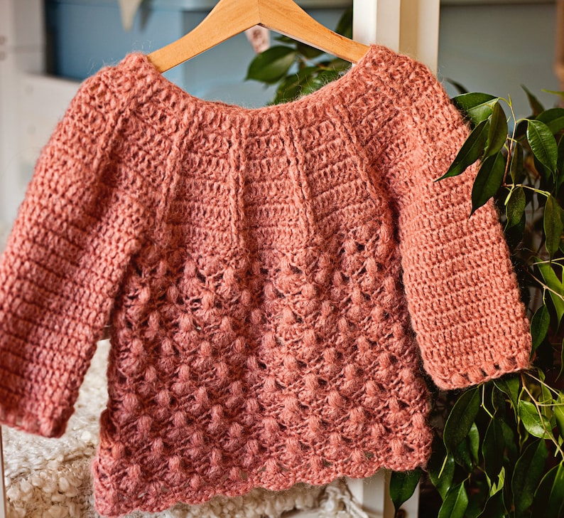 May include: A crocheted sweater in a dusty rose color with a textured stitch pattern. The sweater has a wide neckline and long sleeves.