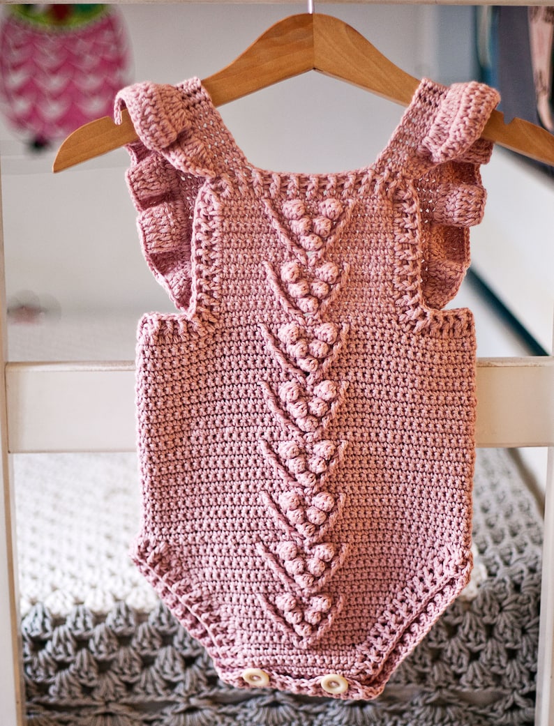 May include: A pink crocheted baby onesie with a ruffled neckline and a textured design. The onesie has a V-shaped pattern with a row of small, raised circles down the center.