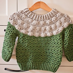 May include: A handmade crochet sweater in green and light gray, displayed on a wooden hanger. The sweater features a textured stitch pattern and a round neckline. The sleeves are long, and the garment appears to be sized for a child.