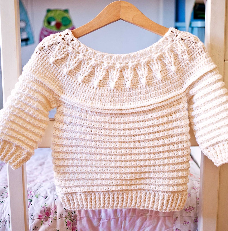 May include: Cream-colored crochet sweater with a round neckline and long sleeves. The sweater features a textured, ribbed design on the body and sleeves, with a decorative pattern around the neckline. It is displayed on a wooden hanger.