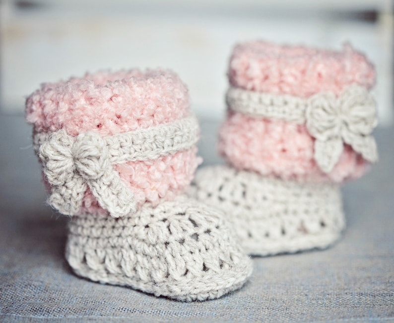 Puede incluir: Un par de botines de beb&eacute; tejidos a crochet con hilo rosa y gris. Los botines tienen una parte superior de color rosa esponjosa y una suela de crochet gris. Cada bot&iacute;n tiene un lazo de crochet en la parte superior.