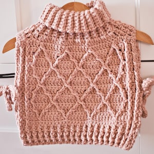 Pode incluir: Um colete de tricô rosa com gola alta e padrão de losangos. O colete tem um pescoço alto e um caimento solto.