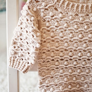 May include: A beige crocheted sweater with a textured, bobble stitch pattern. The sweater has a round neckline and long sleeves.