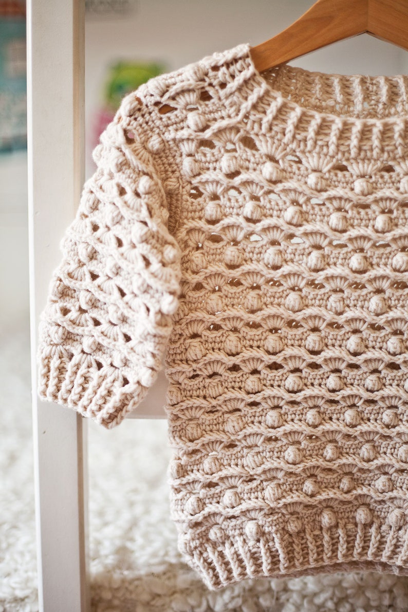 May include: A beige crocheted sweater with a textured, bobble stitch pattern. The sweater has a round neckline and long sleeves.