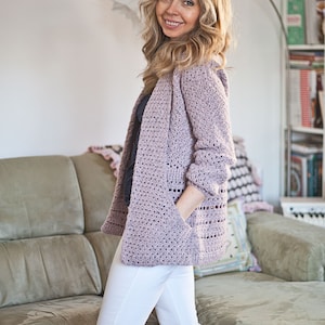 May include: A light purple crocheted cardigan with long sleeves and pockets. The cardigan is open in the front and has a loose fit.