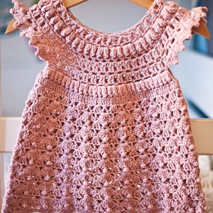 Peut inclure: Une robe de b&eacute;b&eacute; en crochet rose avec un ourlet festonn&eacute; et un motif floral d&eacute;licat. La robe a des manches courtes et un col rond.