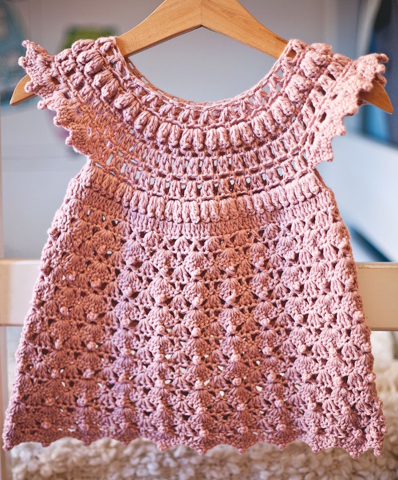 Peut inclure: Une robe de b&eacute;b&eacute; en crochet rose avec un ourlet festonn&eacute; et un motif floral d&eacute;licat. La robe a des manches courtes et un col rond.
